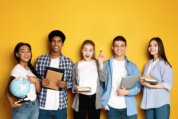 A cheerful, multiethnic group of students stands against a bright yellow backdrop, each holding...