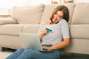 A cheerful young woman is seated on the floor leaning against a couch in a well-lit living room. She is holding a credit card and looking at her laptop screen, likely making an online purchase.