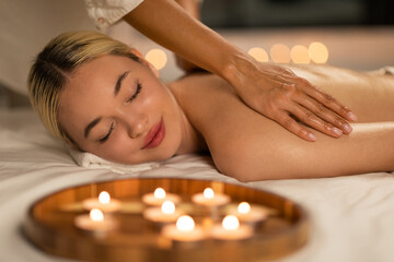 A blonde woman lies face down while enjoying a calming back massage at a spa, her expression one of...