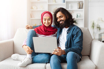 A Muslim couple smiles as they browse the internet on a laptop, seated comfortably in their...