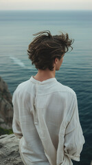 Fototapeta premium young man wearing a loose flowing white shirt or casual attire stands confidently on a cliff overlooking the ocean