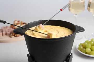 Dipping ham into fondue pot with tasty melted cheese at white table, closeup