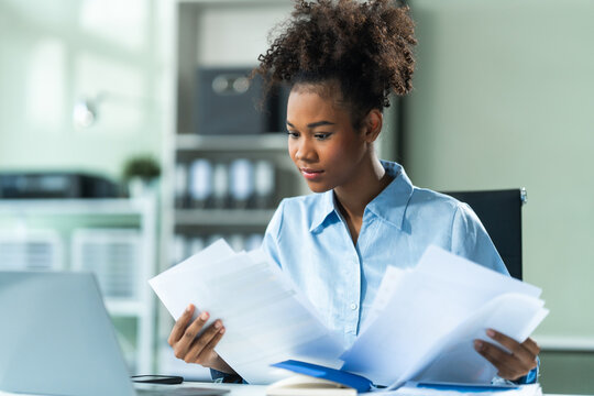 Analyzing business charts A4, a young African American woman in a blue formal shirt and afro brown hair works as a Market Research Analyst in a modern office.