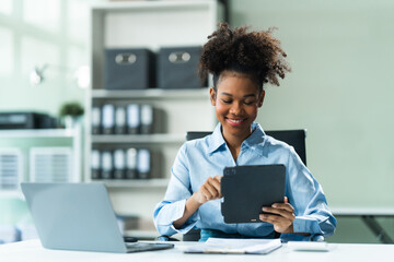 Utilizing a tablet, a young African American woman in a blue formal shirt with afro brown hair works as a Market Research Analyst in a modern office.