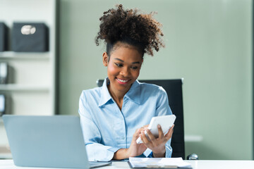 Performing calculations with a calculator, a young African American woman in a blue formal shirt with afro brown hair works as a Market Research Analyst in a modern office.