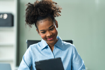 Utilizing a tablet, a young African American woman in a blue formal shirt with afro brown hair...