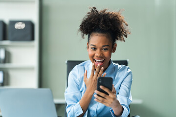 Exuberant and triumphant, a young African American woman in a blue formal shirt with afro brown hair operates a tablet, notebook, and mobile phone in a modern office setting.