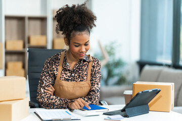 A young African American woman with afro brown hair works in a modern office, managing her small business, utilizing laptop technology for e-commerce, shipping, and marketing, sme box.
