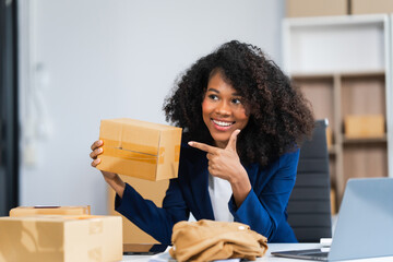 A young African American woman with afro brown hair works in a modern office, managing her online clothing store and live stream platform, sme box packages.