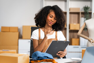 A young African American woman with afro brown hair works in a modern office, managing her online clothing store and live stream platform, sme box packages.
