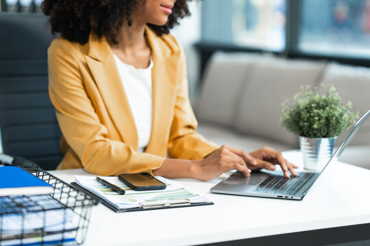 Performing calculations with a calculator, a young African American woman in yellow formal suit with afro brown hair works as a Market Research Analyst in a modern office.