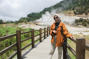 A young woman stands on a wooden bridge leading to Sikidang Crater with white steam.A young woman stands on a wooden bridge leading to Sikidang Crater with white steam. - Powered by Adobe