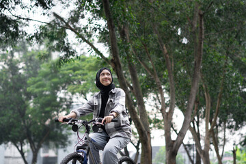 Young cheerful hijab woman, wearing casual flannel riding bicycle bike on sidewalk in city enjoying being outdoors, look aside. People active urban healthy lifestyle cycling concept.