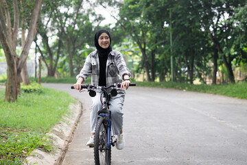 Young cheerful hijab woman, wearing casual flannel riding bicycle bike on sidewalk in city enjoying being outdoors, look aside. People active urban healthy lifestyle cycling concept.