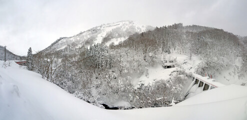 Beautiful Japanese satoyama winter scenery with forest oh the hill covered with snow, mountain river with dam and distant mountains in Ginzan onsen, wide panoramic view