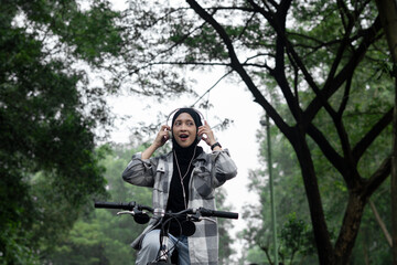 Young cheerful hijab woman, wearing casual flannel riding bicycle bike on sidewalk in the city park enjoying being outdoors, look aside or ahead. People active urban healthy lifestyle cycling concept.