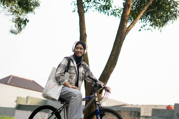 Young cheerful hijab woman, wearing casual flannel riding bicycle bike on sidewalk in the city park enjoying being outdoors, look aside or ahead. People active urban healthy lifestyle cycling concept.