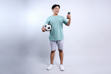 Full Length Of Young Asian Man Holding Soccer Ball And Looking At Smartphone With Smilling Face...