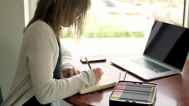 creative professional engrossed in designing a logo, using traditional tools like colored pencils and a sketchbook, with a laptop open beside her for digital enhancements. The setting highlights