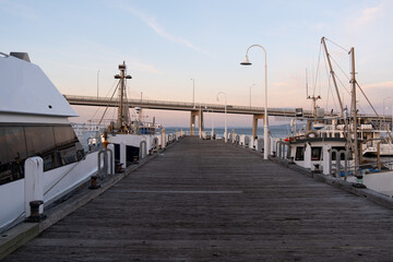 Wooden boardwalk at San Remo Jetty with ships berthed and Phillip Island bridge in the distance during sunset. Background texture of a timber pier at the shore with copy space.