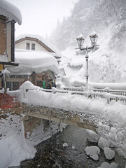 idyllic Japanese small village winter scenery with traditional houses, big snowdrifts on the roofs, wooden bridge, river, forest in snow on the hill, Ginzan onsen hot spring, Yamagata, Japan