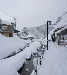 Walking in Beautiful Japanese small village street in winter with traditional houses, big snow drifts, river, pine forest in snow on the hill, Ginzan onsen hot spring, nature of Yamagata, Japan