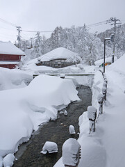 Walking in Beautiful Japanese small village street in winter with traditional houses, big snow drifts, river, pine forest in snow on the hill, Ginzan onsen hot spring, nature of Yamagata, Japan