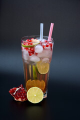 A faceted tall glass of fruit juice with ice and straws on a black background, next to pieces of ripe lime and a pomegranate fruit with seeds.