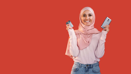Mobile banking. Online shopping. Satisfied cheerful happy woman in hijab showing credit card phone isolated on red empty space background.