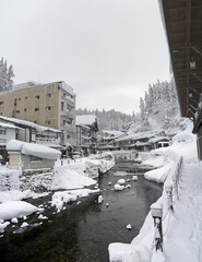 Beautiful Japanese small village winter scenery ith traditional houses with snowdrifts on the roofs and wooden bridges, river, pine forest in snow on the hill, Ginzan onsen hot spring, Yamagata, Japan