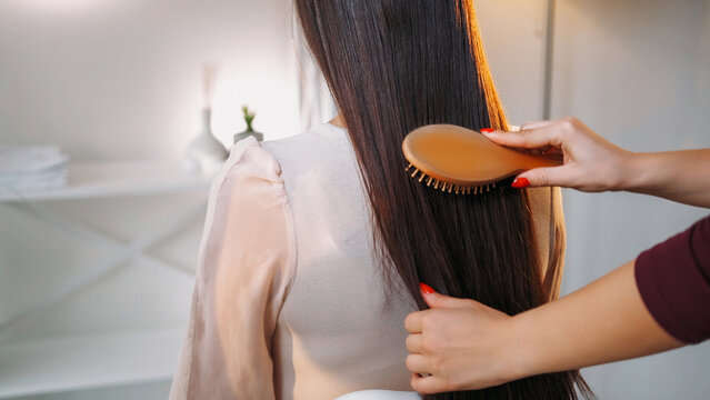 Professional hairstylist. Salon treatment. Woman hands combing model lady beautiful smooth long brunette hair with wooden brush in light interior.