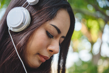 Happy asian woman use headphones listen to music from smartphone outdoor in green park nature....