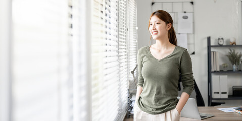 Happy smiling young businesswoman standing in office with copy space. Confident asian woman in casual wear standing in workplace.