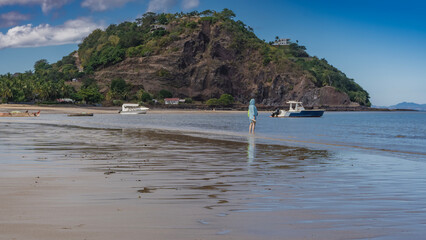 The low tide in the ocean. Puddles on the sandy beach. A man stands at the edge of the water, looking into the distance. The boats are anchored to the shore. A hill against a blue sky and clouds.