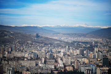 expansive aerial view of Tbilisi, Georgia, showcasing a dense urban landscape with various buildings under a clear blue sky, bordered by mountains in the distance