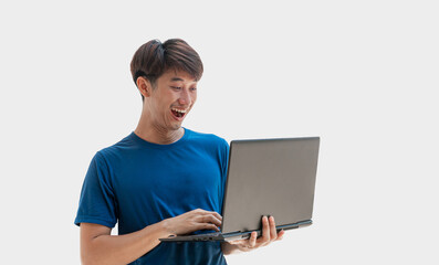 A young Asian man in his 20s wearing a blue t-shirt using a laptop for online remote work happily isolated on a gray background. Man watching webinars on a laptop, e-learning, e-banking, Looks funny