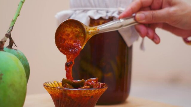 Close up shot of Homemade Mango Pickle or aam ka achar in bowl with raw mangoes and transparent jar in background. Indian homemade raw mango pickle. Indian woman hand with spoon of pickle. 