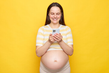 Joyful satisfied beautiful young pregnant woman wearing striped top posing isolated over yellow...
