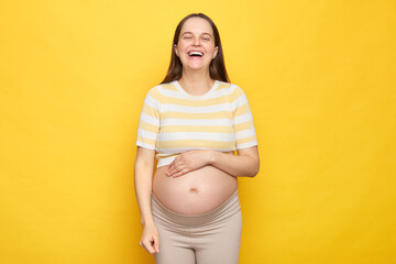 Happy funny brown haired pregnant woman with bare belly posing isolated over yellow background...
