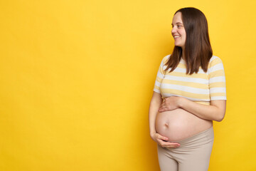 Cheerful delighted brown haired pregnant woman with bare belly posing isolated over yellow...