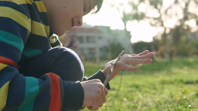 Side view, tight shot of caucasian cute boy, removing soil form his hand, using a wooden stick. Blurry fields on the backround slow motion 120fps