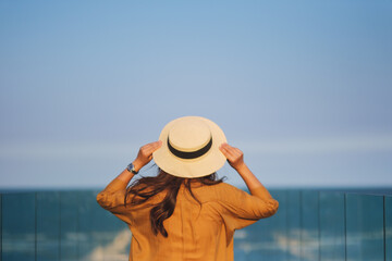 Naklejka premium Rear view image of a woman with hat standing on glass terrace with sea and blue sky background