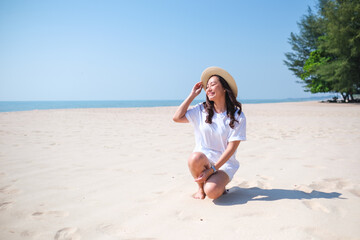 Portrait image of a young woman with white t-shirt and hat sitting on the beach with the sea and blue sky background