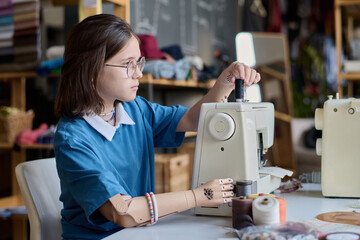 Side view portrait of teenage girl with prosthetic hand using sewing machine in vocational training...