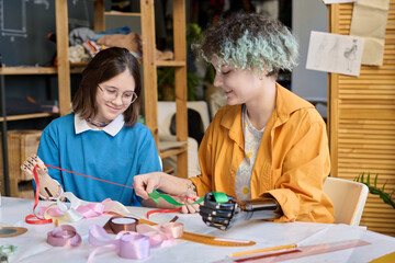 Portrait of two teenage girls enjoying sewing class in inclusive training center and holding...