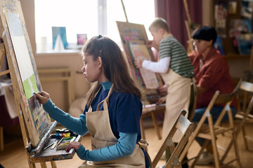 Side view at children with disabilities enjoying art therapy class and painting on easels with...