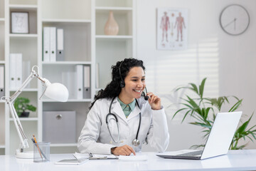 A cheerful Hispanic female doctor in conversation on the phone while sitting at her office desk,...
