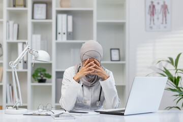 A female doctor in a hijab sits at her office desk, visibly stressed and overwhelmed, with her head...