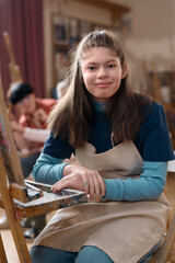 Vertical portrait of smiling young girl with disability sitting by easel in art class and looking...
