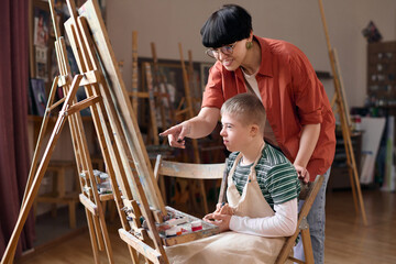 Side view portrait of smiling female teacher assisting boy with down syndrome painting on easel in...